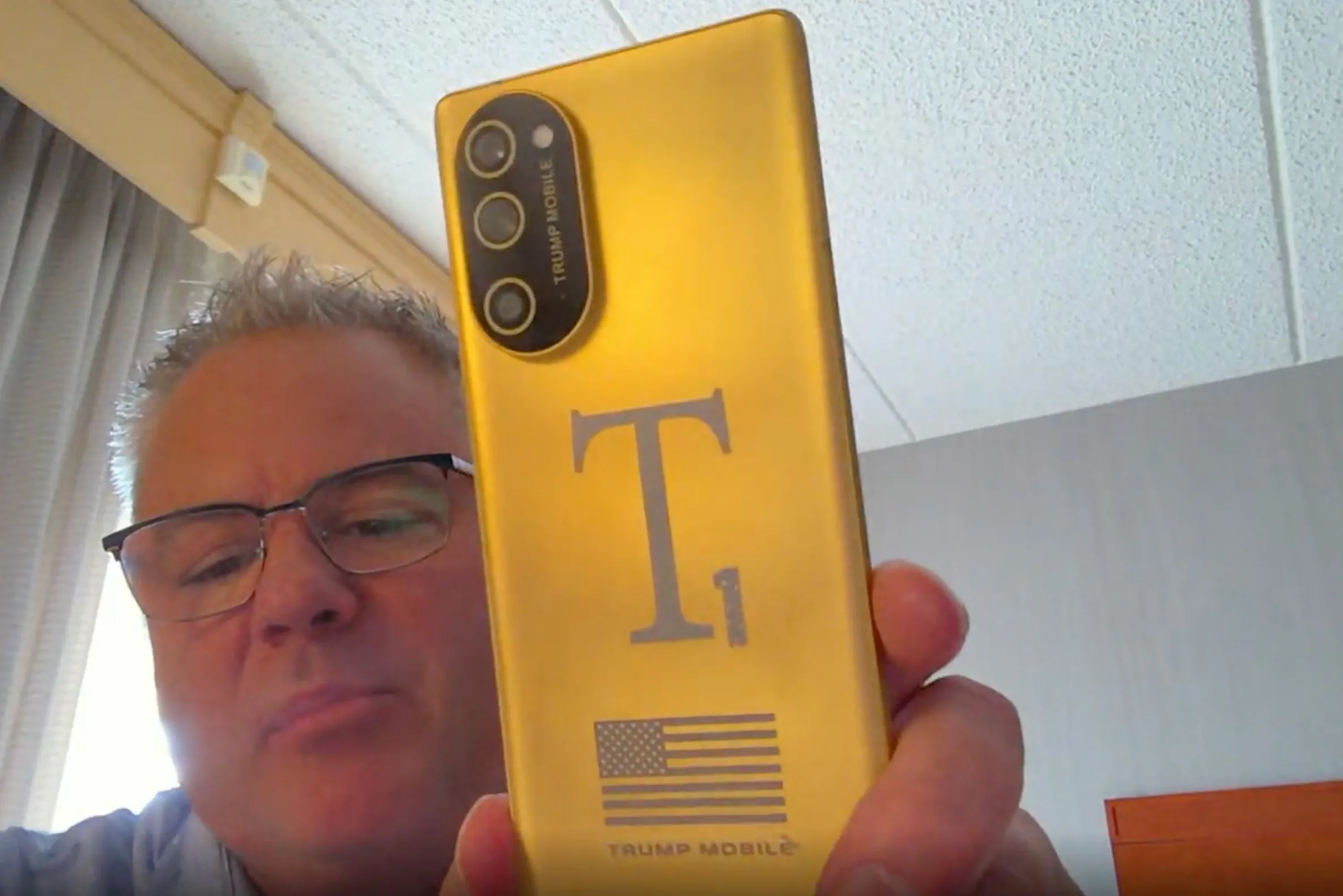 Screenshot of Eric Thomas from Trump Mobile holding a gold T1 Phone towards his webcam
