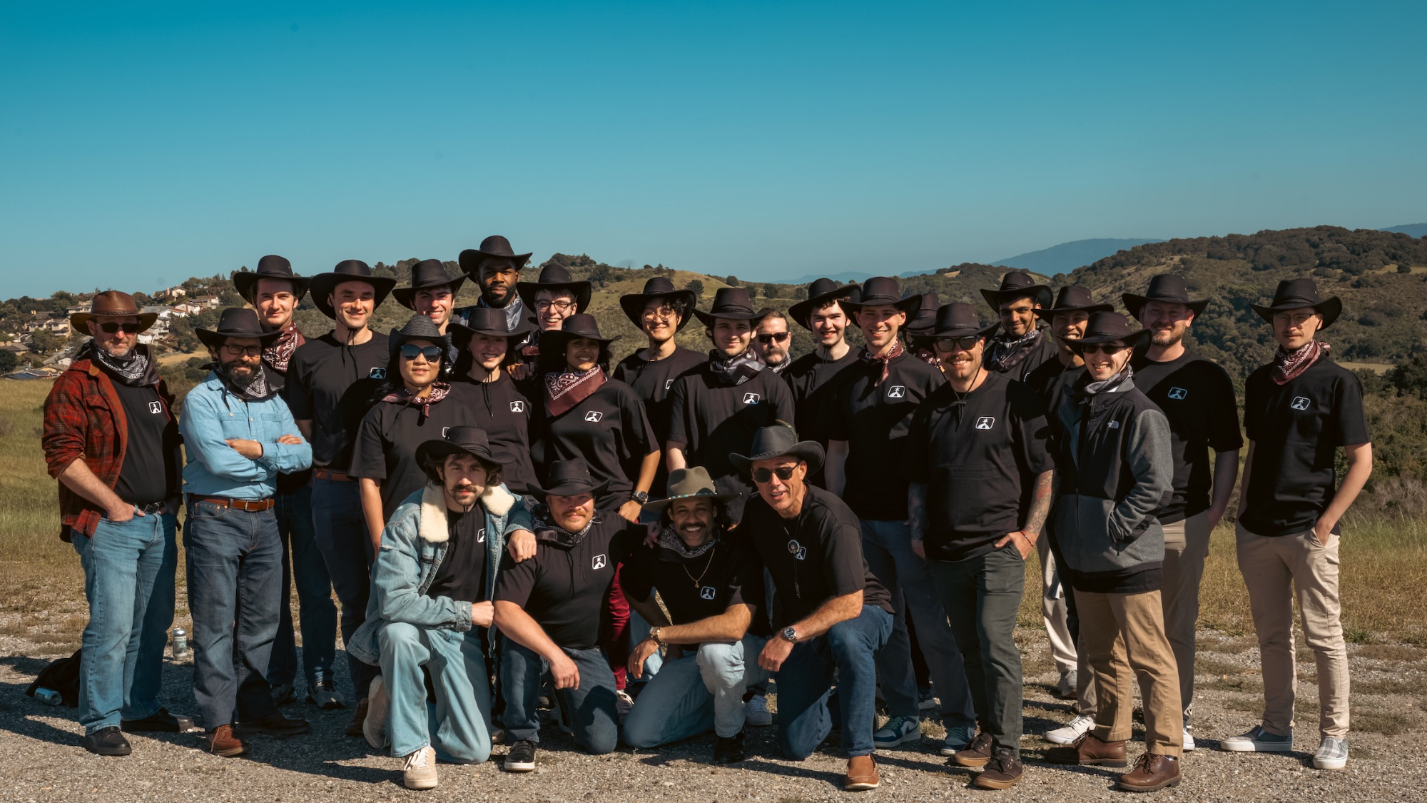 several dozen people wearing black shirts and black cowboy hats standing together outside with sparsely forested hills in the background