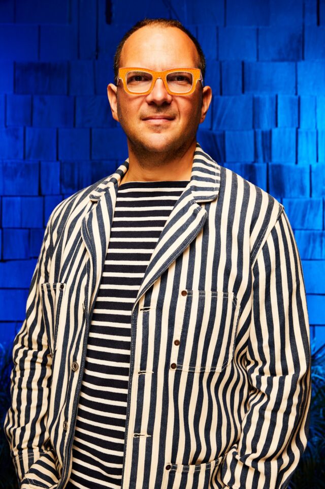 Balding man wearing striped shirt and jacket standing against blue background with hands in pockets.
